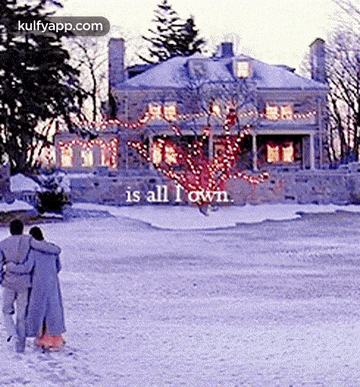 a man and woman are walking in the snow in front of a large house ..