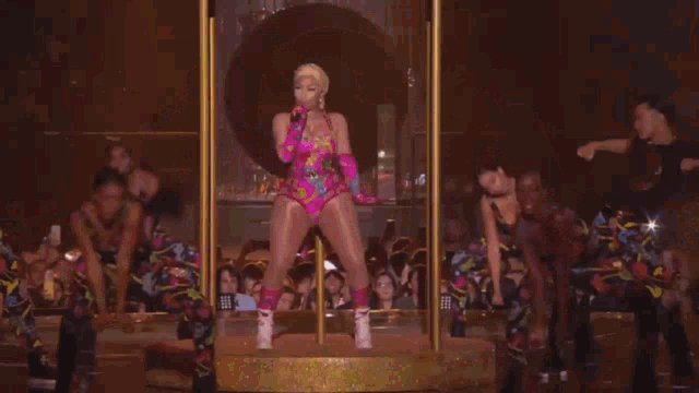 a woman in a pink bodysuit is standing on a stage with a group of dancers .