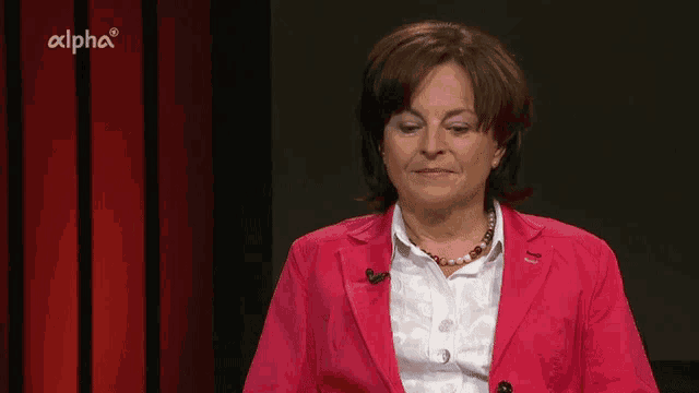a woman in a pink jacket and white shirt is sitting in front of a red curtain with alpha written on it