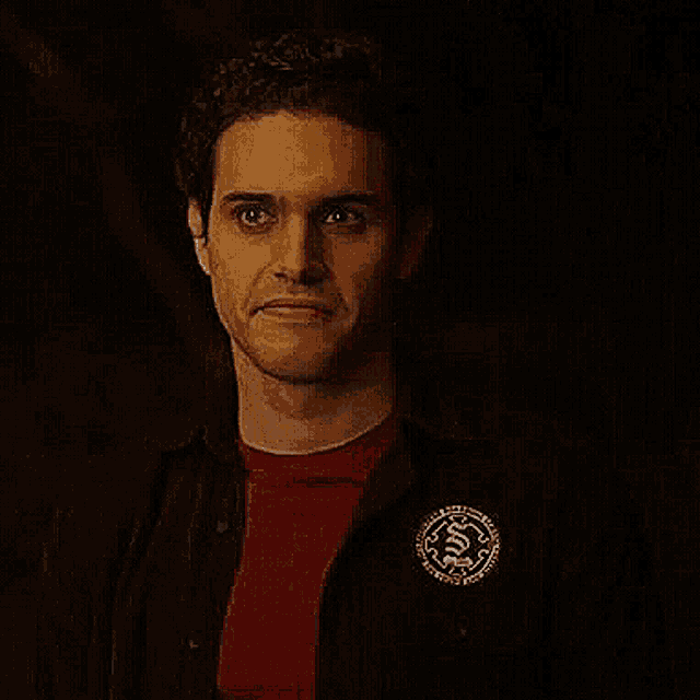 a man wearing a red shirt and a black jacket with a pin on his chest is looking at the camera .