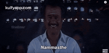 a man in a white shirt is smiling and saying namasthe in front of a crowd .