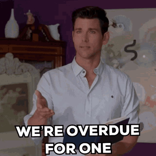 a man in a white shirt holds a book and says we 're overdue for one