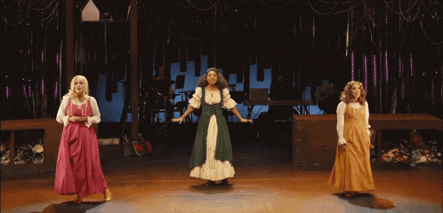 three women in long dresses are singing on a stage in front of a keyboard