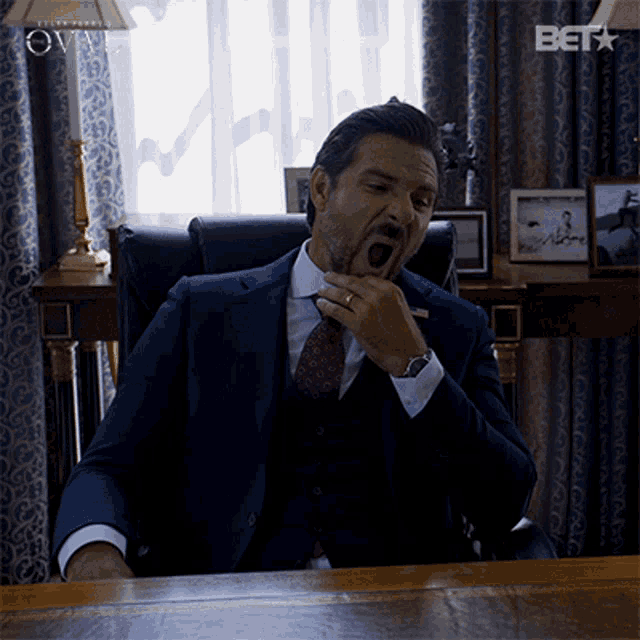 a man in a suit and tie is yawning while sitting at a desk with a bet logo in the corner