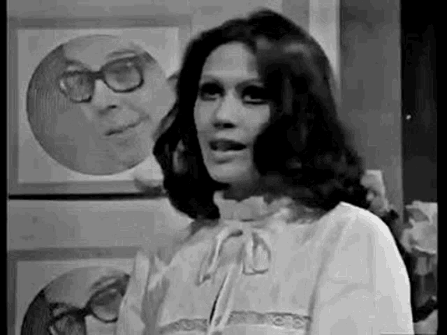 a black and white photo of a woman standing in front of a mirror with a picture of a man in the background .