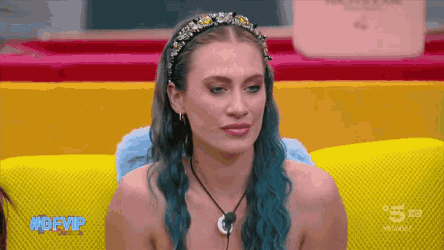 a woman with blue hair is sitting on a yellow couch in front of a sign that says #gfvp