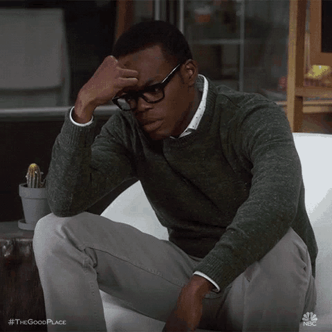 a man wearing glasses and a green sweater sits on a couch with his hand on his forehead