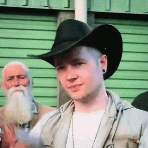 a man wearing a cowboy hat and a vest stands in front of a green wall