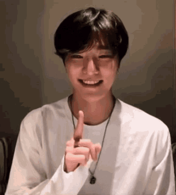 a young man wearing a white shirt and a necklace is smiling and pointing at something .