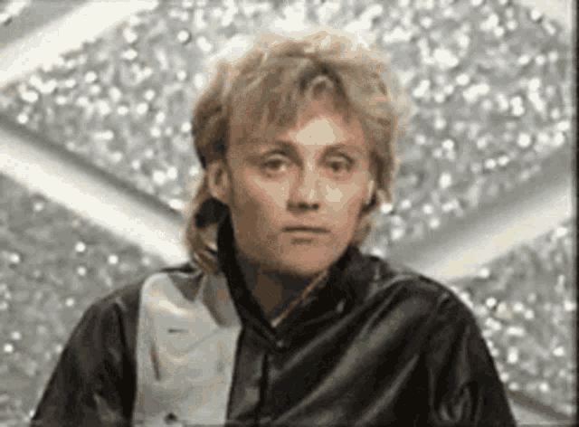 a man with a mullet is wearing a black leather jacket and looking at the camera .