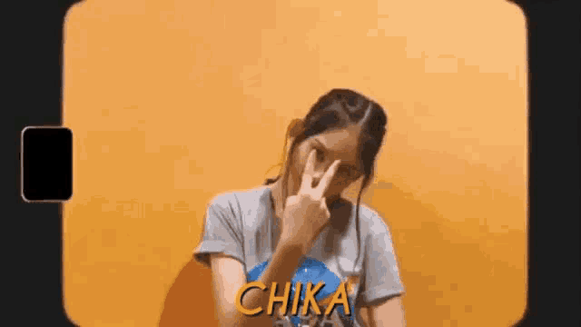 a woman is making a peace sign in front of a yellow wall with the word chika on it .