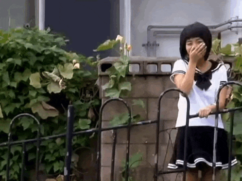 a girl in a school uniform is standing next to a fence covering her mouth .