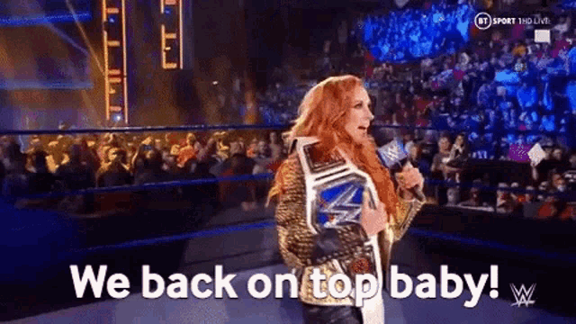 a woman in a wrestling ring holding a championship belt says we back on top baby .