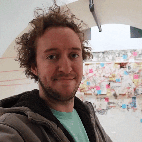 a man with curly hair and a beard smiles in front of a wall with sticky notes on it