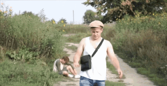 a man is walking down a dirt road while another man sits on the grass