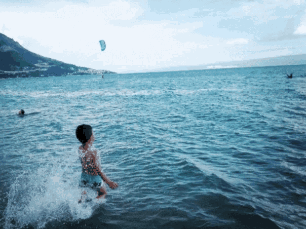 a boy is splashing in the ocean with a kite flying in the background