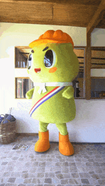 a green mascot with a yellow hat and orange boots is standing on a tiled floor