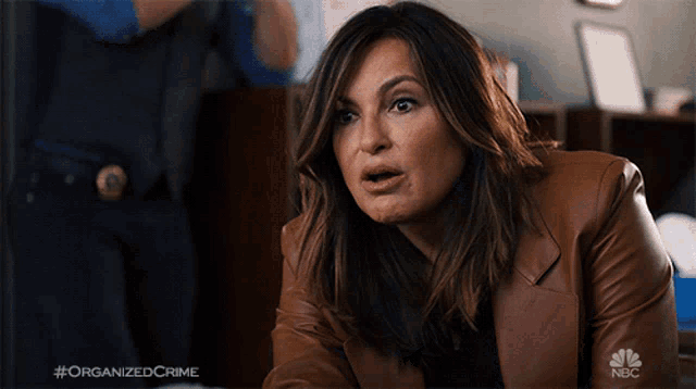 a woman in a brown leather jacket is sitting in front of a nbc sign