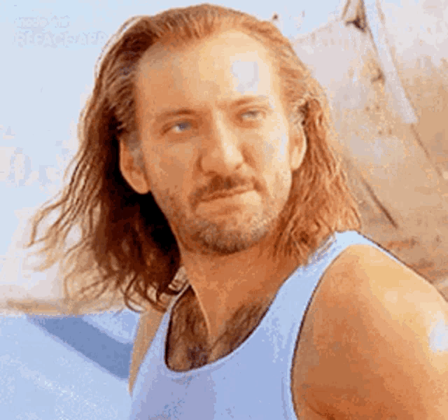 a man with long hair and a beard wearing a blue tank top is looking at the camera .