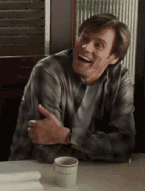 a man in a plaid shirt is laughing while sitting at a table with a cup of coffee