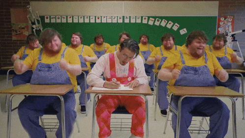 a group of men in overalls are sitting at desks in a classroom with a green board behind them