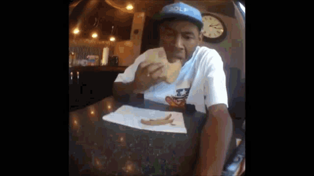 a man is sitting at a table eating a sandwich with a clock on the wall behind him .