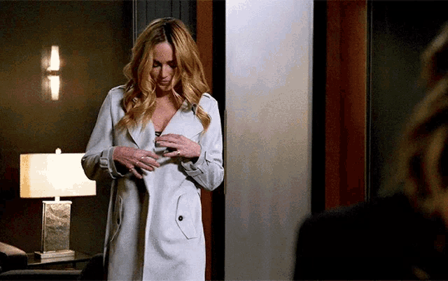 a woman wearing a white trench coat is standing in a room with a lamp