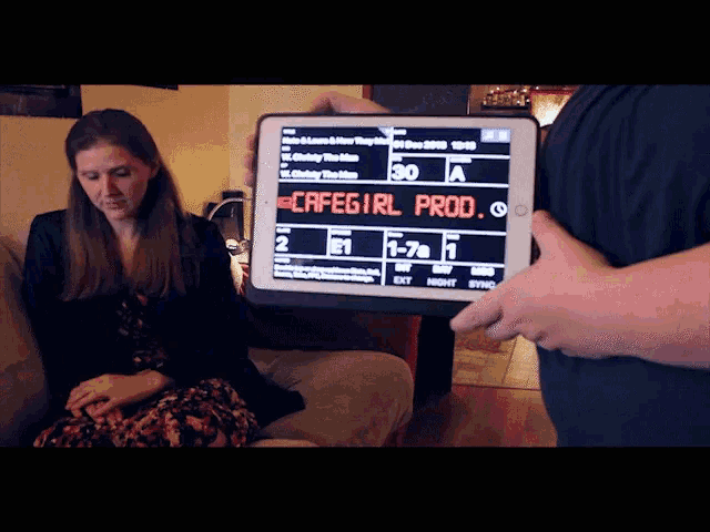 a woman sits on a couch while a man holds up a tablet that says cafegirl prod