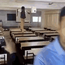 a man in a blue shirt is standing in an empty classroom with tables and chairs .
