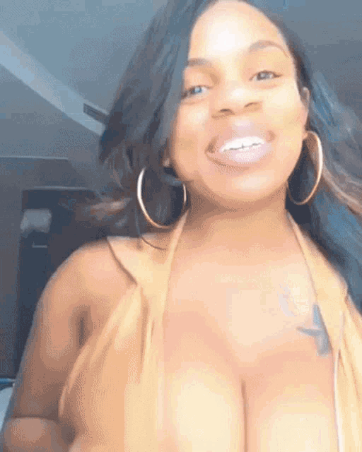 a woman with a tattoo on her chest is smiling while wearing a yellow top and hoop earrings .