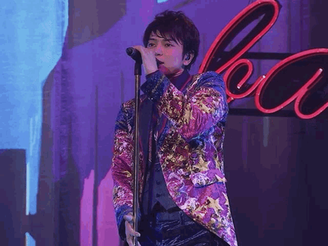 a man in a floral jacket sings into a microphone in front of a neon sign that says coca cola