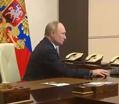 a man in a suit sits at a desk with a flag in the background