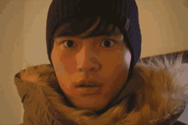 a young man wearing a beanie and a fur hooded jacket looks surprised