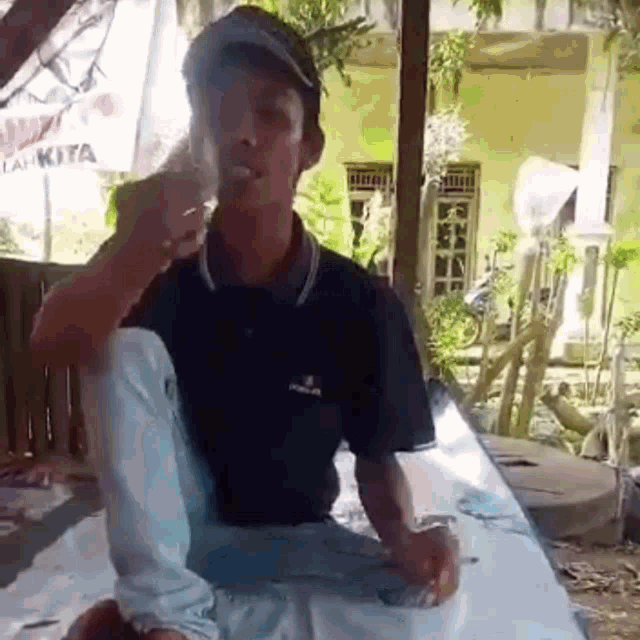 a man is sitting at a table smoking a cigarette while wearing a baseball cap .