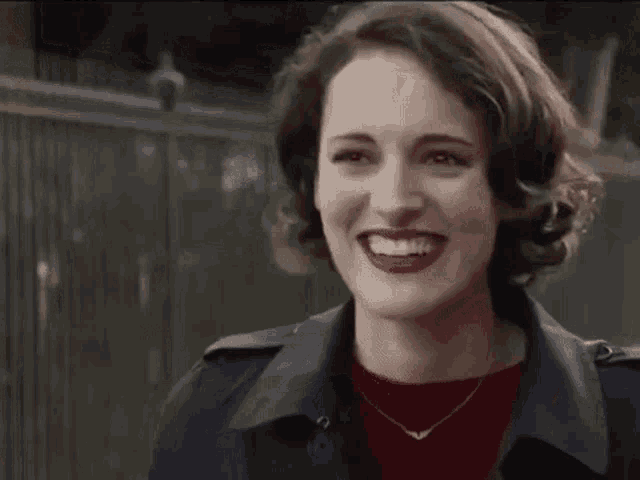 a woman in a trench coat and red sweater is smiling and looking at the camera .