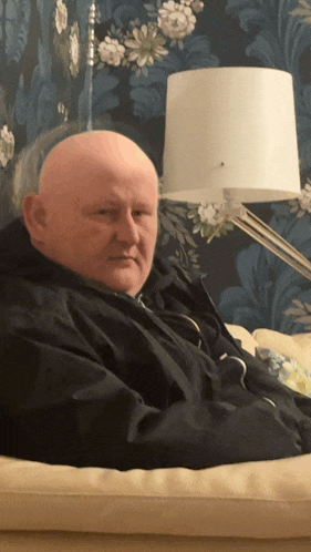 a bald man is sitting on a couch with a lamp in the background