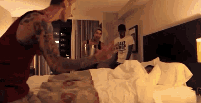 a man wearing a shirt that says " cake shop " stands next to a man laying in bed