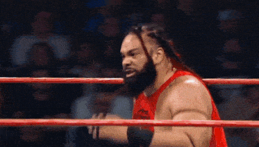 a wrestler in a red tank top is standing in a ring .