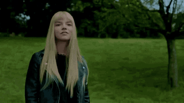 a woman with blonde hair is standing in a field wearing a black leather jacket .