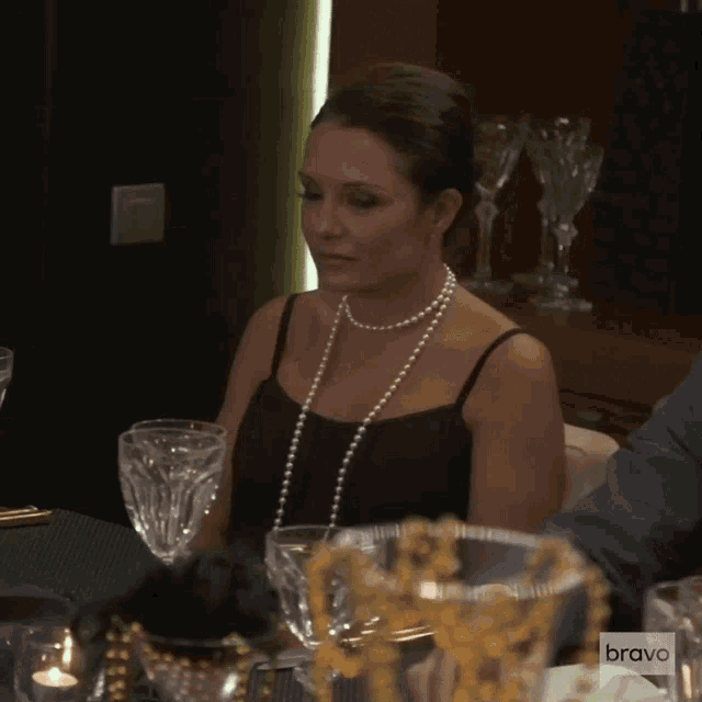 a woman wearing a pearl necklace sits at a table with a bravo logo in the foreground