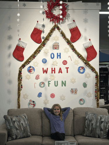 a little girl is sitting on a couch in front of a wall that says oh what fun