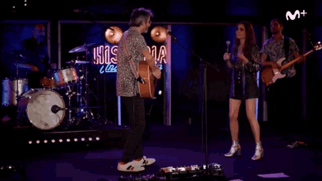 a man playing an acoustic guitar stands next to a woman singing in front of a sign that says m +