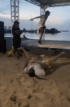 a man is laying on the ground on the beach while another man jumps in the air .