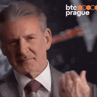 a man in a suit and tie applauds in front of a btc prague logo