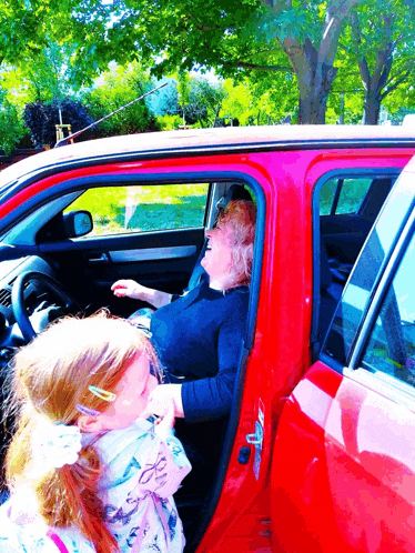 an older woman sits in a red car while a little girl looks out the window