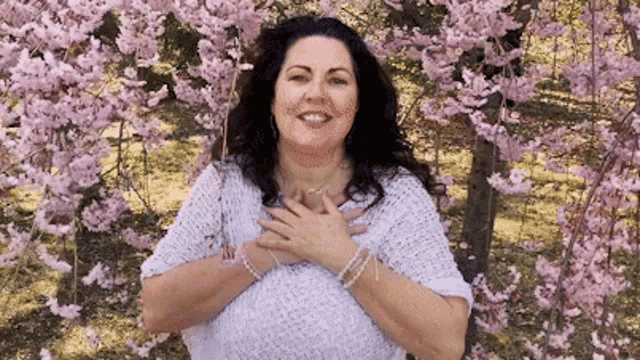a woman is standing in front of a cherry blossom tree with her hands on her chest and smiling .