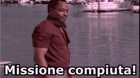 a man in a red shirt is standing in front of a body of water and says missione compiuta !