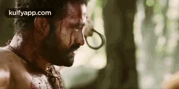 a man with blood on his face is standing in the woods with a ring around his neck .