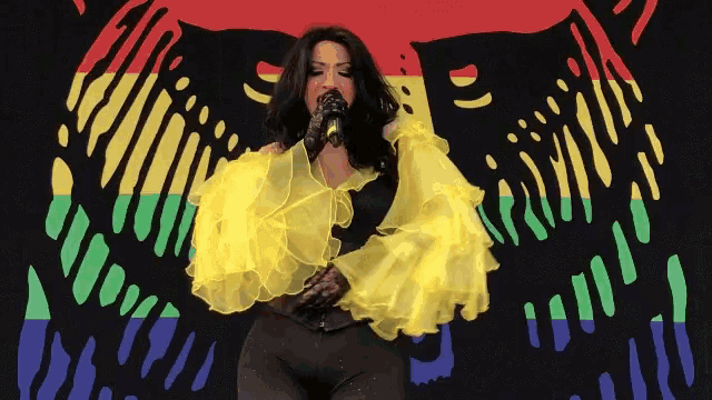 a woman in a yellow ruffled top sings into a microphone in front of a colorful background