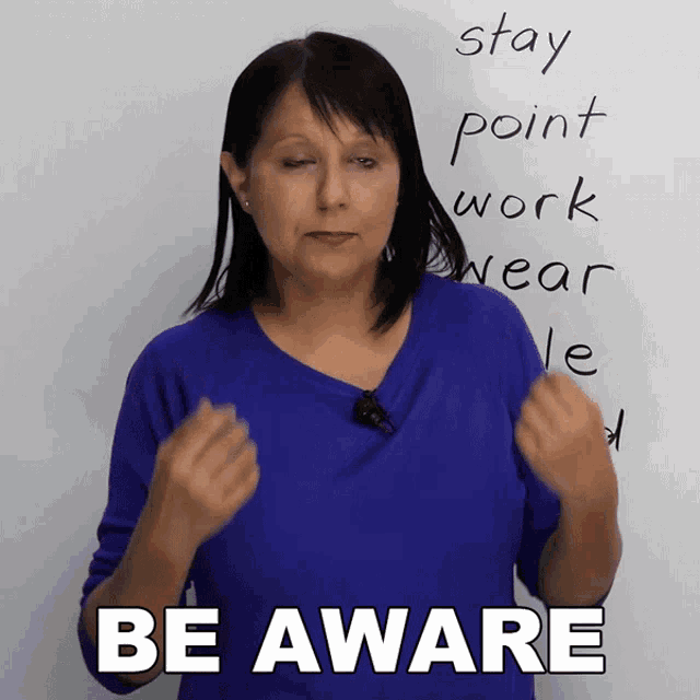 a woman in a blue shirt says be aware in front of a whiteboard